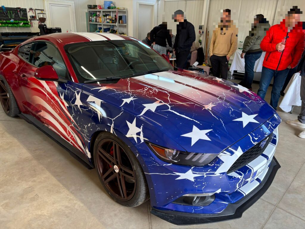 Ford Mustang colantat integral cu design steag american, fetele persoanelor din fundal sunt blurate pentru confidentialitate